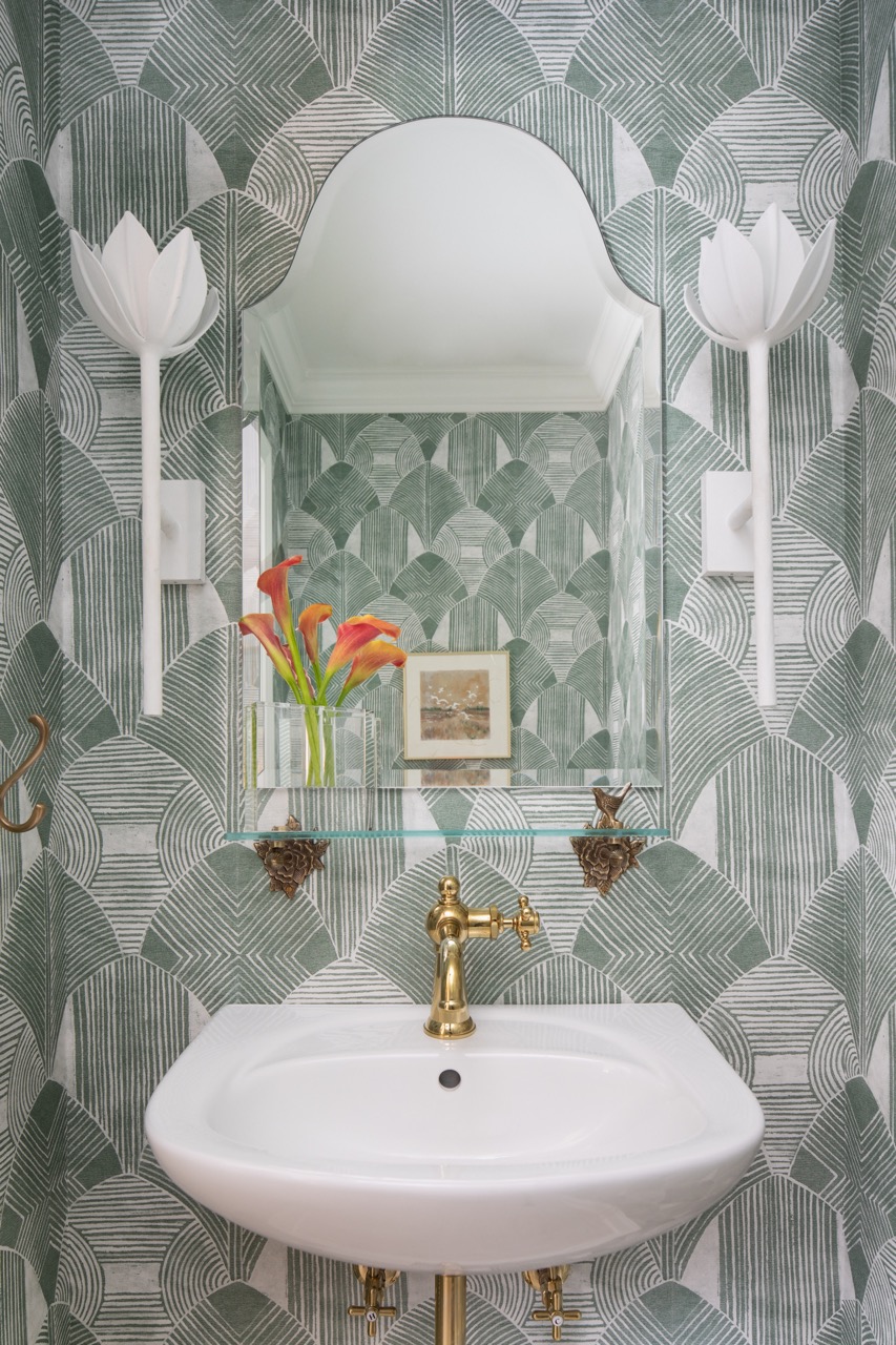 A sink with a patterned wallpaper behind it and a mirror above it sits against a wall with a gold faucet. 