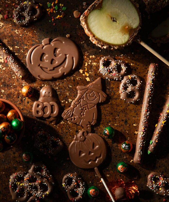 Where to Buy Local Halloween Candy in Pittsburgh Various Halloween character shaped chocolates, small candies, chocolate covered pretzels, and a cut-in-half chocolate apple sit on a dark brown background.