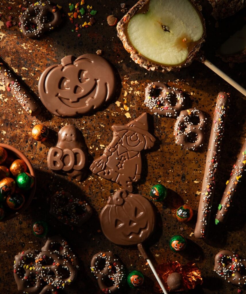 Various Halloween character shaped chocolates, small candies, chocolate covered pretzels, and a cut-in-half chocolate apple sit on a dark brown background.