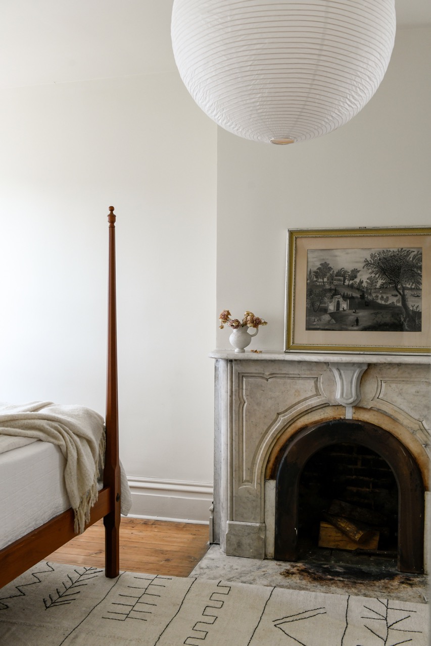 Another picture of a living area of a townhouse featuring a white fireplace with a framed photo on top, bed and brown bed post, and a white lantern light in the center of the ceiling. 