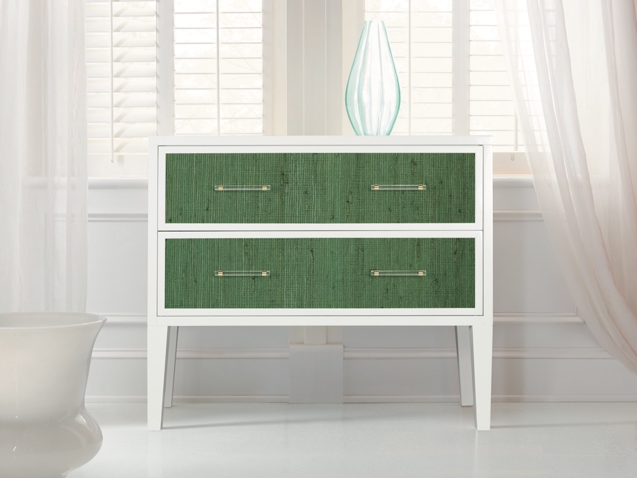 A white dresser with green drawers sits in front of white windows and curtain. 