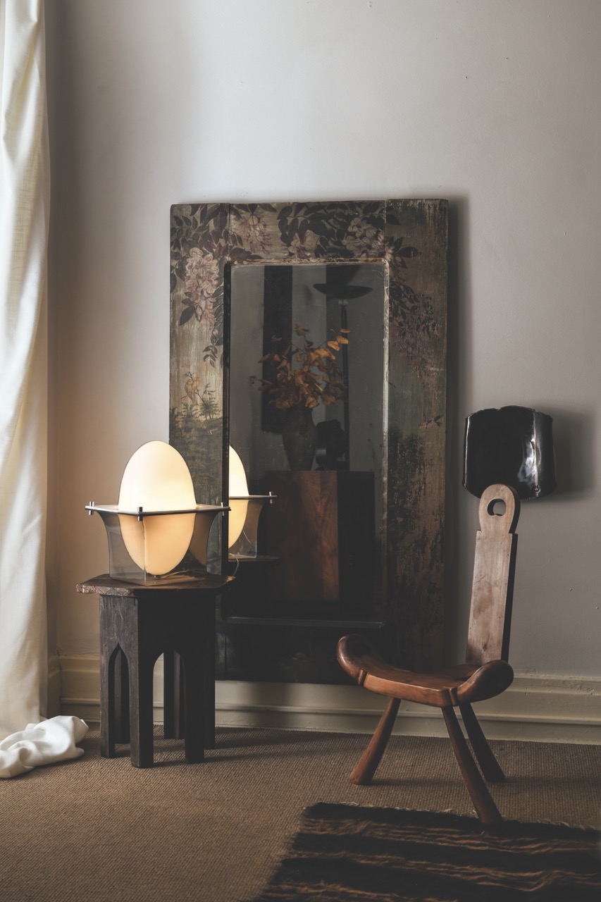 A large mirror stands against a wall behind a tall lamp and vintage wooden chair. 