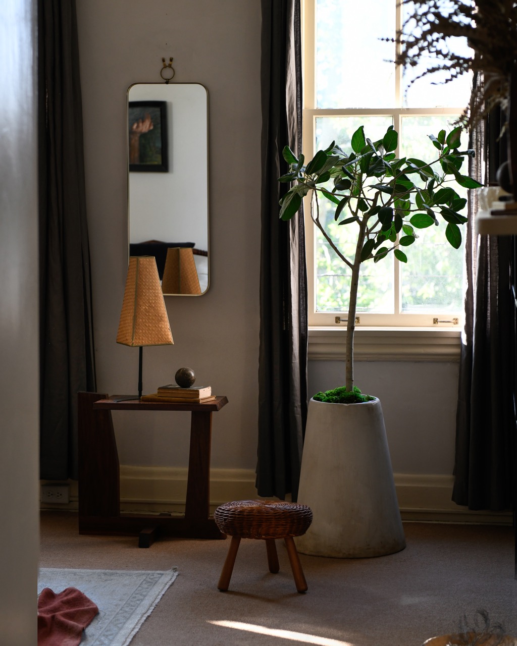 A plant in a vintage vase sits in front of a window with a small table to the left featuring a yellow lamp. 