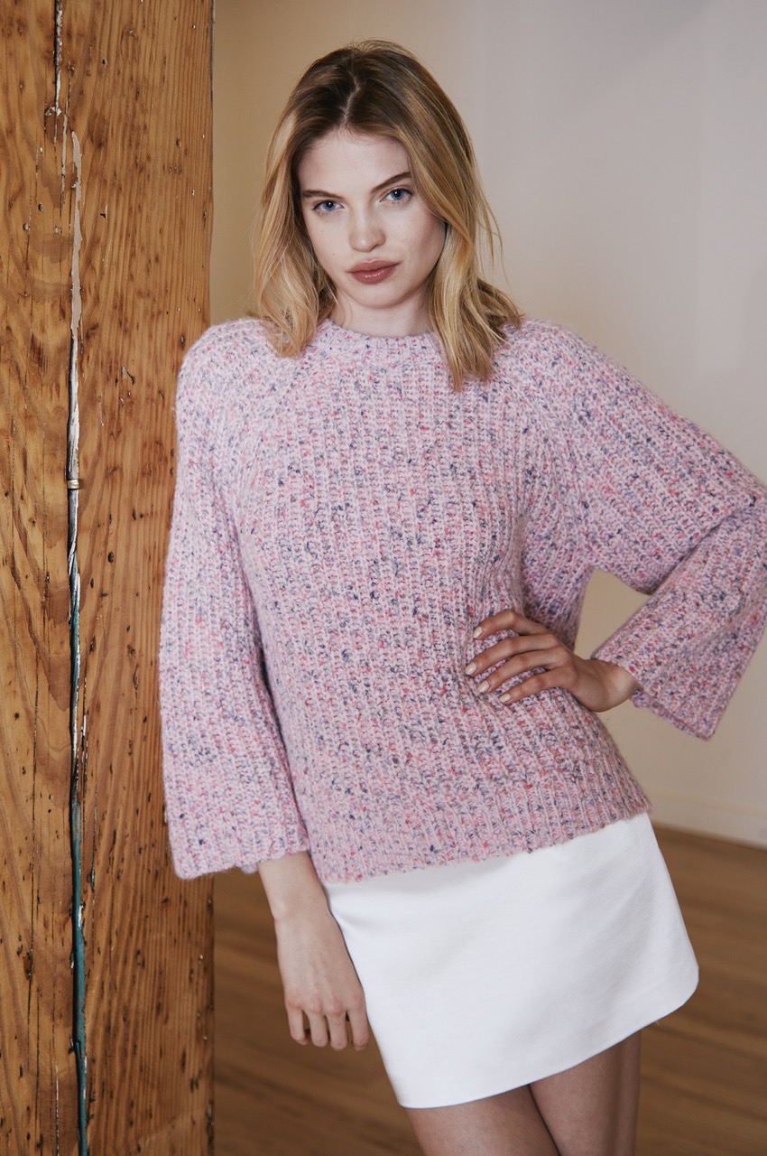 A woman stands in a pink sweater and white skirt, leaning against a wall. 