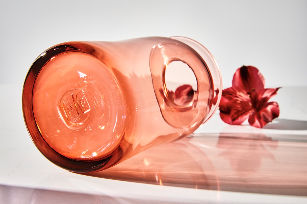 A blush colored glass vase with the same color flower in it lays sideways on a white background.