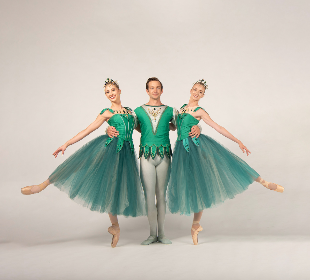 Three ballet dancers, two in long tutus and one in pants, dressed in emerald green stand beside each other