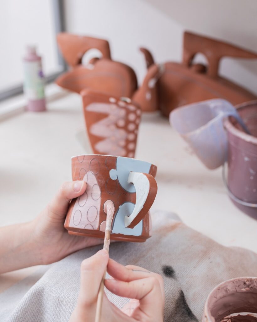 Taylor Mezo Brings a Pop of Color with Her Ceramics A woman paints on a brown ceramic mug.