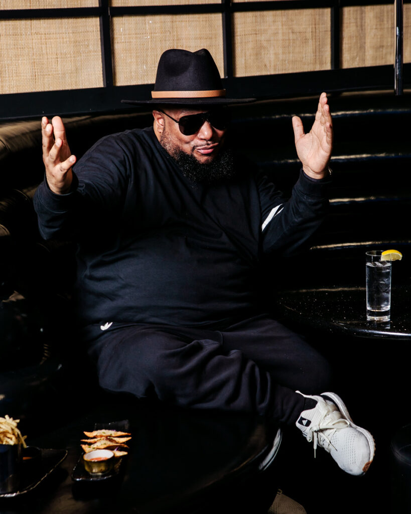 A person seated at a table wearing a black outfit including a hat and sunglasses.