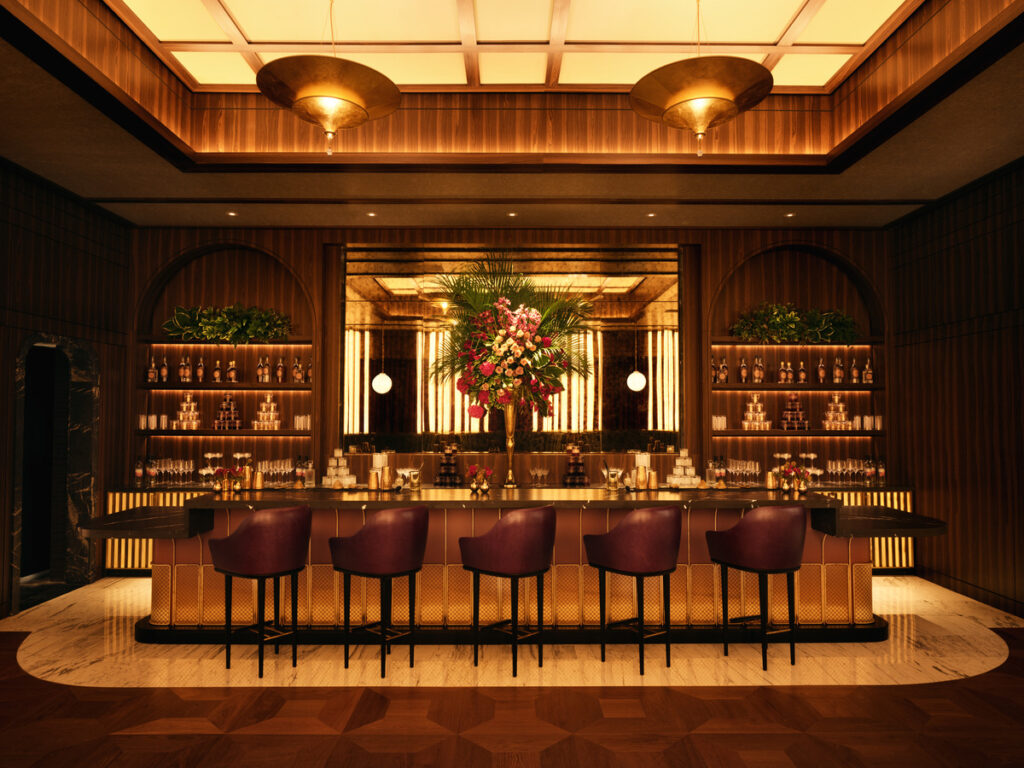 A wood-paneled bar interior with a floral centerpiece on the dark bar counter. Bottle-lined shelves and a crystal chandelier above.