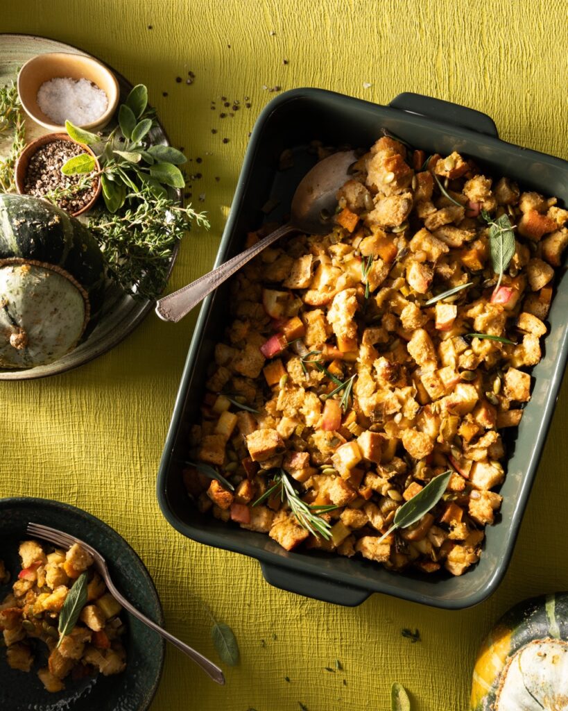 A small rectangular pan of vegan stuffing with a spoon in it sits on a green holiday table.