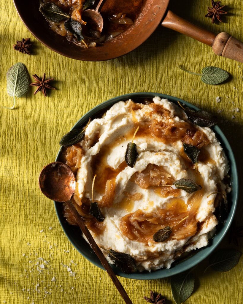 A pot of mashed potatoes mixed with browned butter, sage, and caramelized onions sits on a green table.