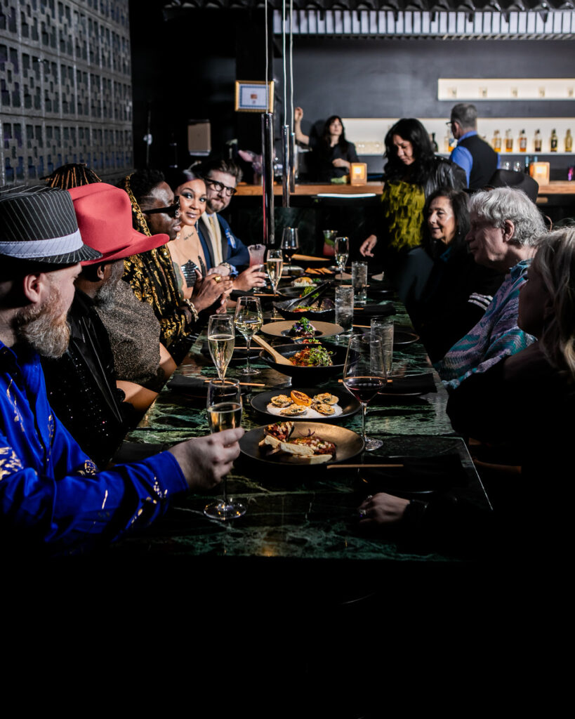 A group of people seated around a long dining table enjoying a meal together. The table is set with plates of food, glasses of wine, and other dining utensils in a warmly lit dining establishment.