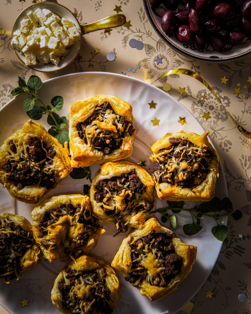 Plate of baked empanadas filled with beef, onion, and cheese mixture topped with feta cheese and served with olives.