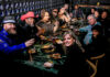 Frzy’s Gratitude Feast A group photo of 12 people sitting around a large dining table surrounded by hanging string lights and potted plants. They are smiling and laughing while holding drinks and conversing with each other.