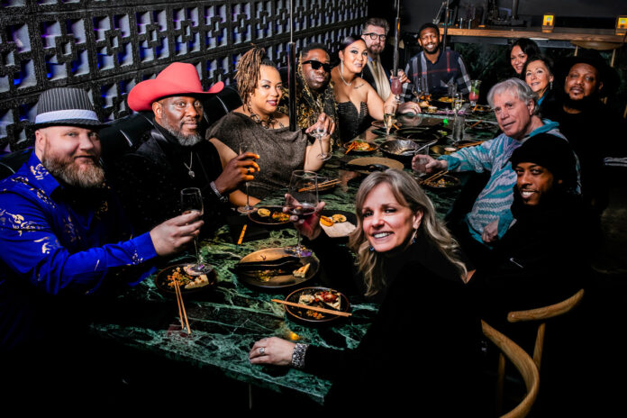A group photo of 12 people sitting around a large dining table surrounded by hanging string lights and potted plants. They are smiling and laughing while holding drinks and conversing with each other.