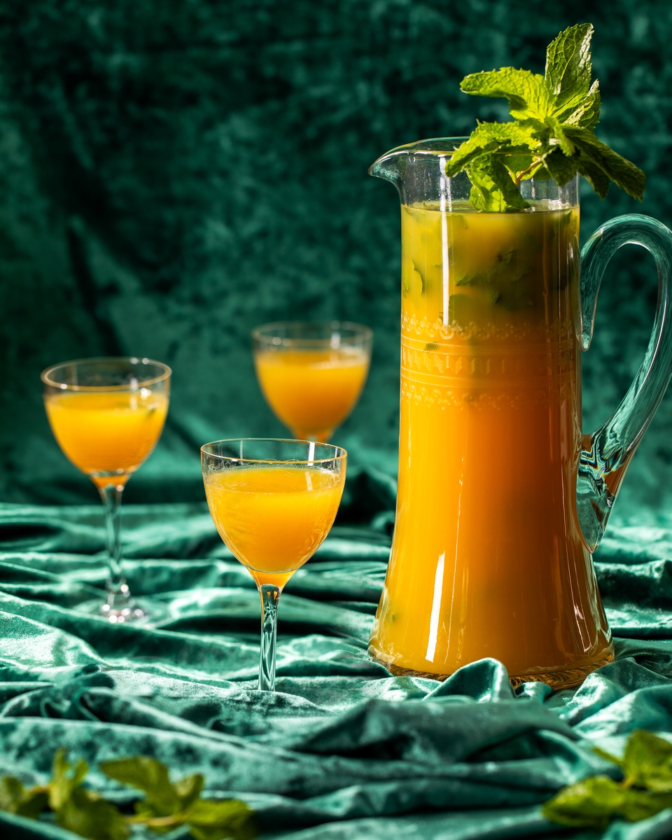 A large glass pitcher filled with a vibrant orange Piña Paradise Mojito cocktail garnished with mint, beside two glasses ready to serve.