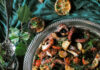 Grilled Octopus with Warm Potato Salad A silver serving dish filled with grilled octopus tentacles, potato wedges, and a garnish of fresh herbs from a tomato and olive salad presented on a green cloth setting.