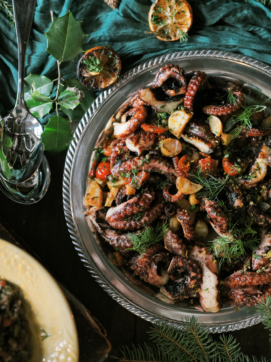 A silver serving dish filled with grilled octopus tentacles, potato wedges, and a garnish of fresh herbs from a tomato and olive salad presented on a green cloth setting.