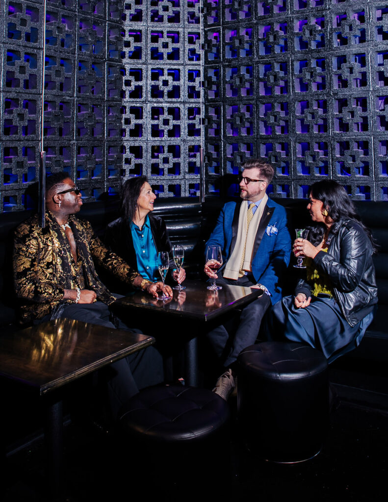 Three people sitting around a table with drinks, in a room with a geometric patterned wall in the background.