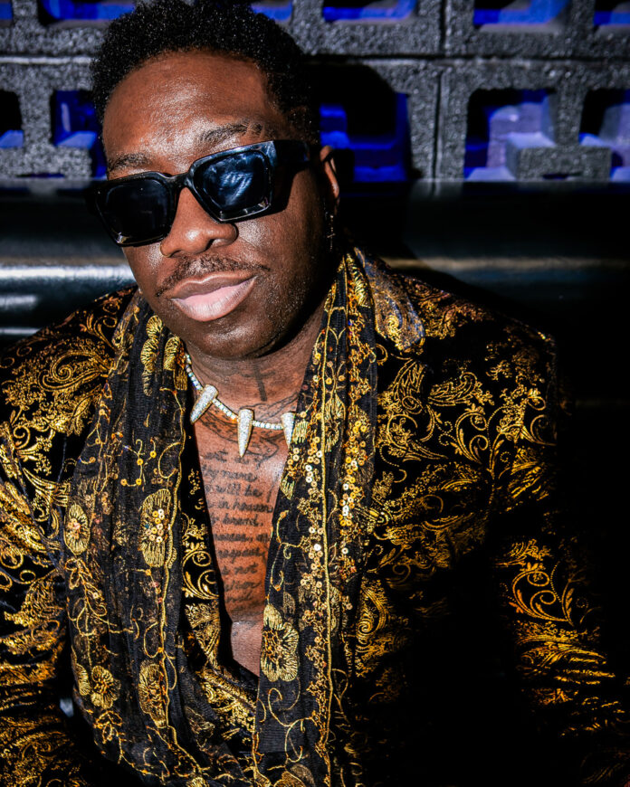 Frzy stands in a yellow shirt and white bone necklace/