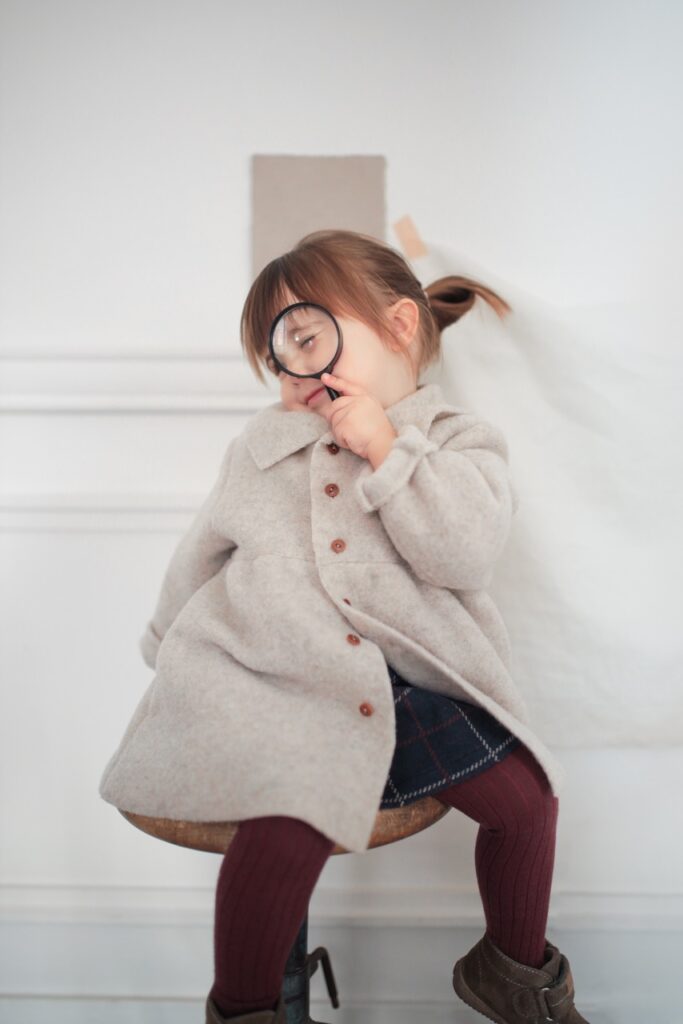 A little girl in a grey jacket holds up a magnifying glass to her eye.