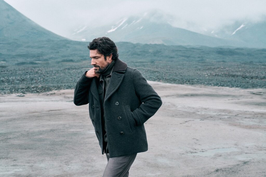 A man walks around in a fleece jacket with mountains behind him.