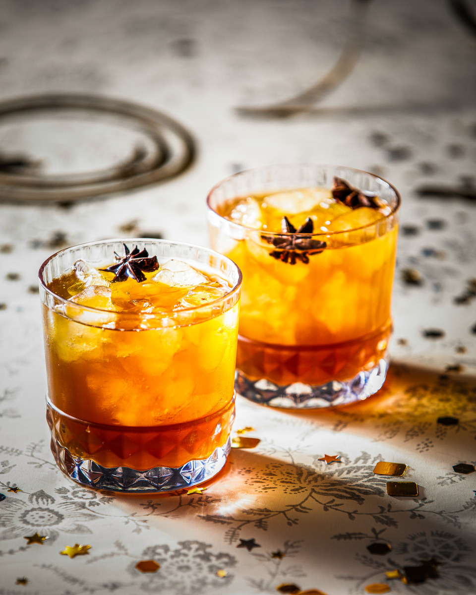 Two glasses of orange-colored mulled cider or similar festive spiced beverage topped with star anise, placed on a patterned tablecloth.