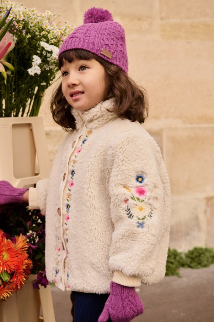 A little girl in a wool jacket with flower embroidery and a purple winter hat.