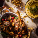 Lamb shanks garnished with rosemary and almonds served alongside rice and nut sauce on a patterned tablecloth.