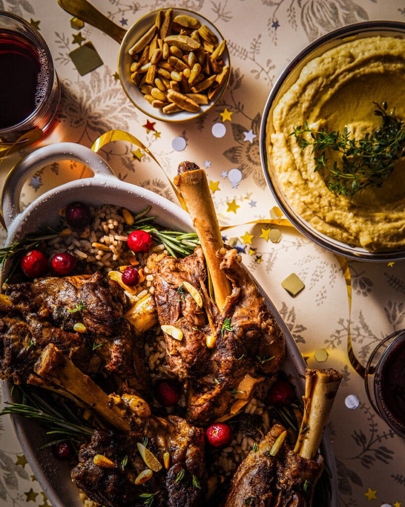 Lamb shanks garnished with rosemary and almonds served alongside rice and nut sauce on a patterned tablecloth.