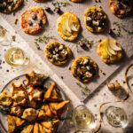 Mini pizzas display featuring pear-apricot-brie, tomato-mozzarella-olive, and pesto-goat cheese-mushroom toppings laid out elegantly on a patterned tablecloth alongside wine glasses and decorations.