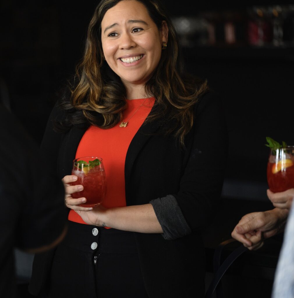 A woman in a red shirt and black jacket smiles with a drink in her hand.