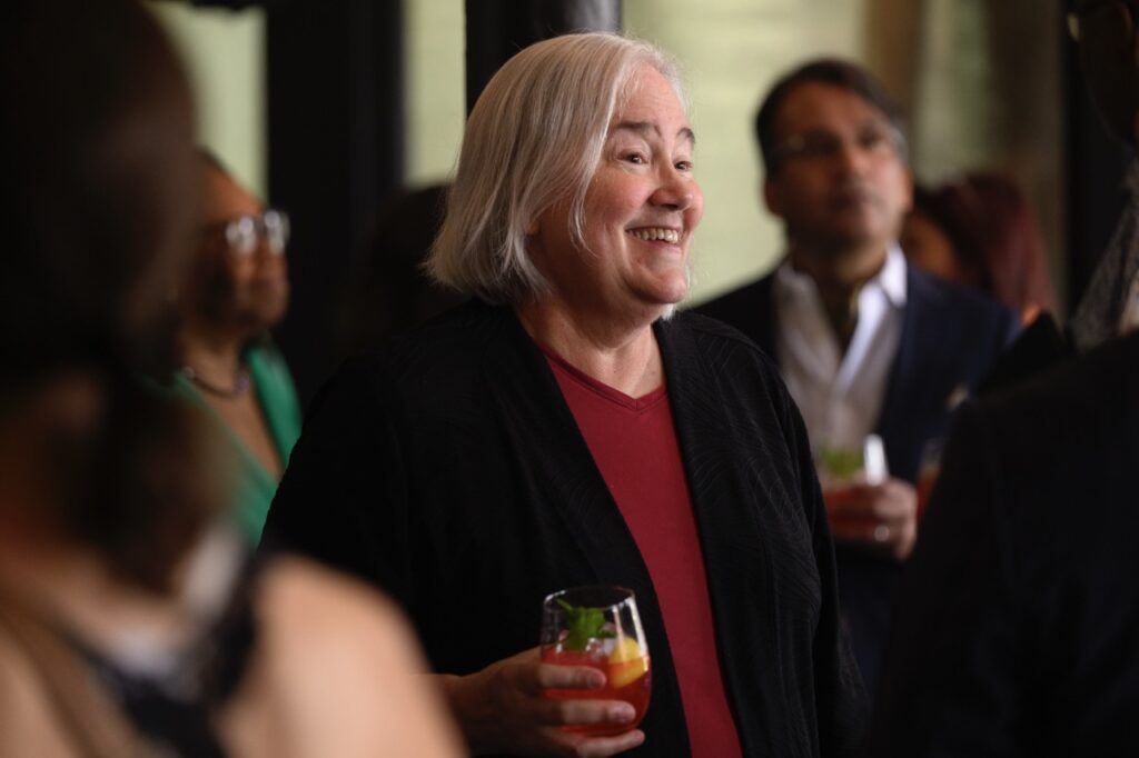 A woman in a red shirt and a black jacket smiles with a cocktail in hand.