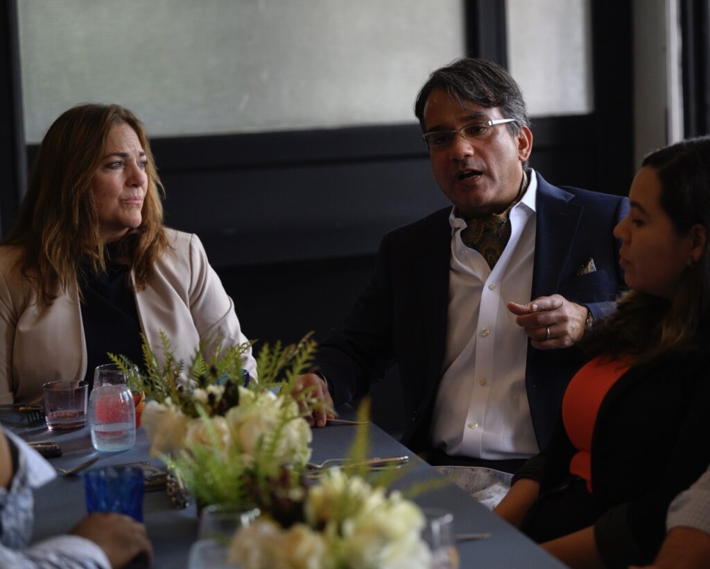 A man in a white button up shirt and suit jacket sits beside a woman in a khaki coat at the table.