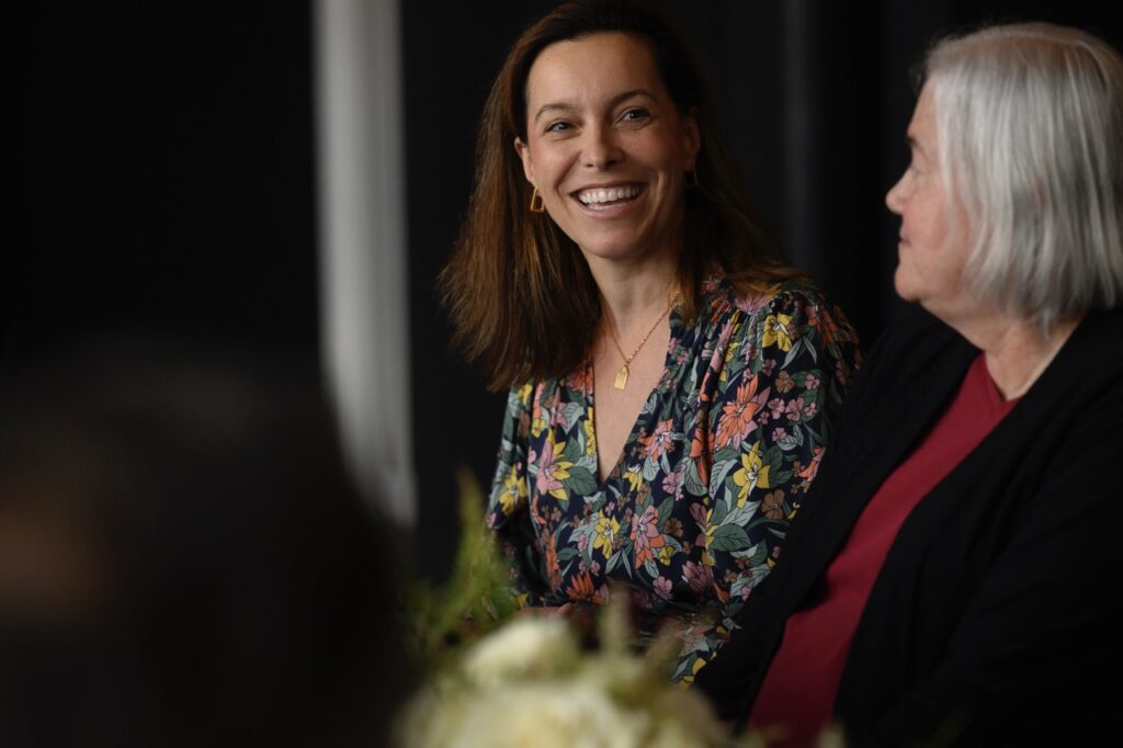 A woman in a floral blouse smiles at a woman in a red shirt and black jacket at a nonprofit roundtable.