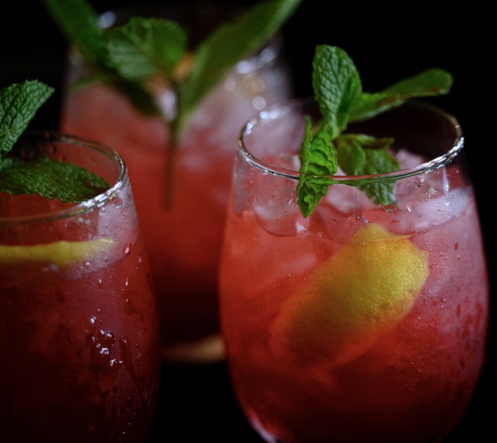 Three glasses with red juice, a mint sprig, and lemon slices sit in a trio together.