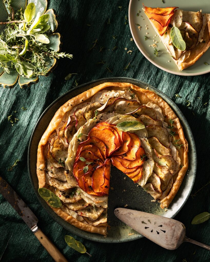 A round root vegetable galette with vegetables spiraled on top sits on a green plate with a slice taken out of it.