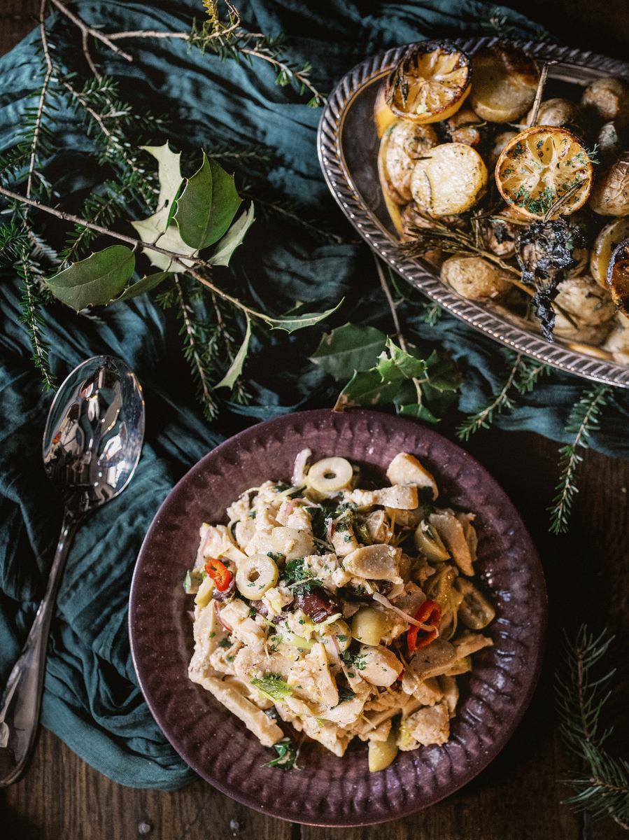 A festive meal setup featuring a bowl of pinkish-white sliced conch salad with lemon, fennel and olive garnishes, accompanied by roasted fingerling potatoes and serving utensils.