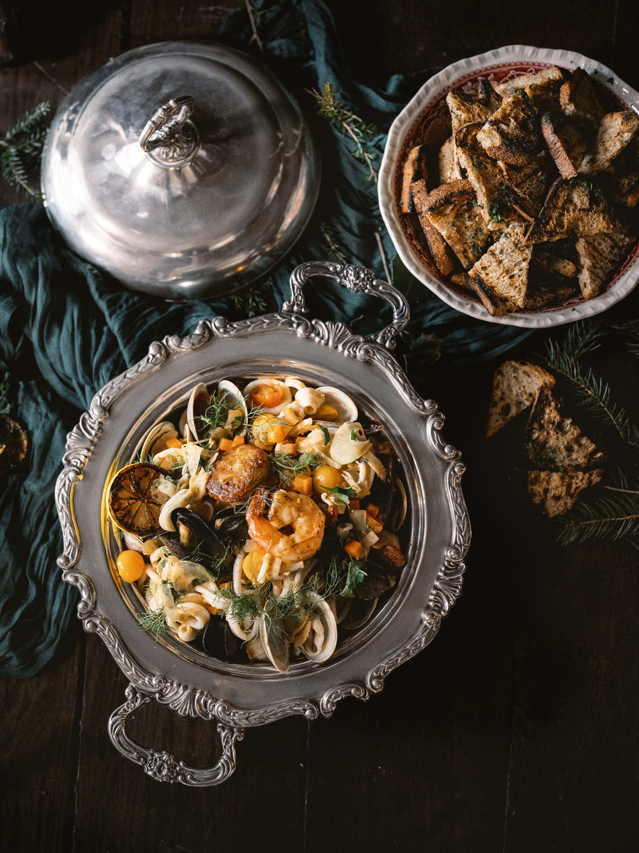 A white serving platter filled with various types of cooked seafood including shrimp, scallops, mussels and calamari rings in a creamy sauce of caramelized fennel, pancetta and white wine.