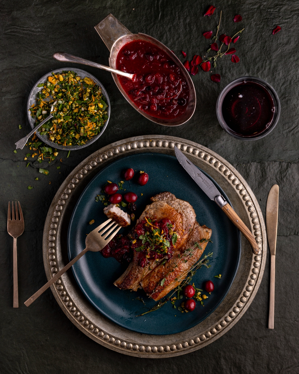 A plate with a seared pork chop topped with cranberry-bourbon sauce, alongside small bowls of shaved Brussels sprouts salad.