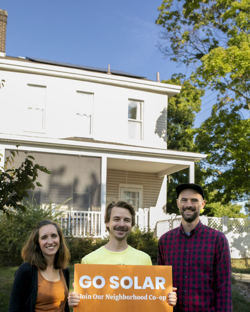 Three individuals from Solar Neighbors United hold a sign that reads "GO SOLAR - Join Our Neighborhood Co-op" in front of a house.