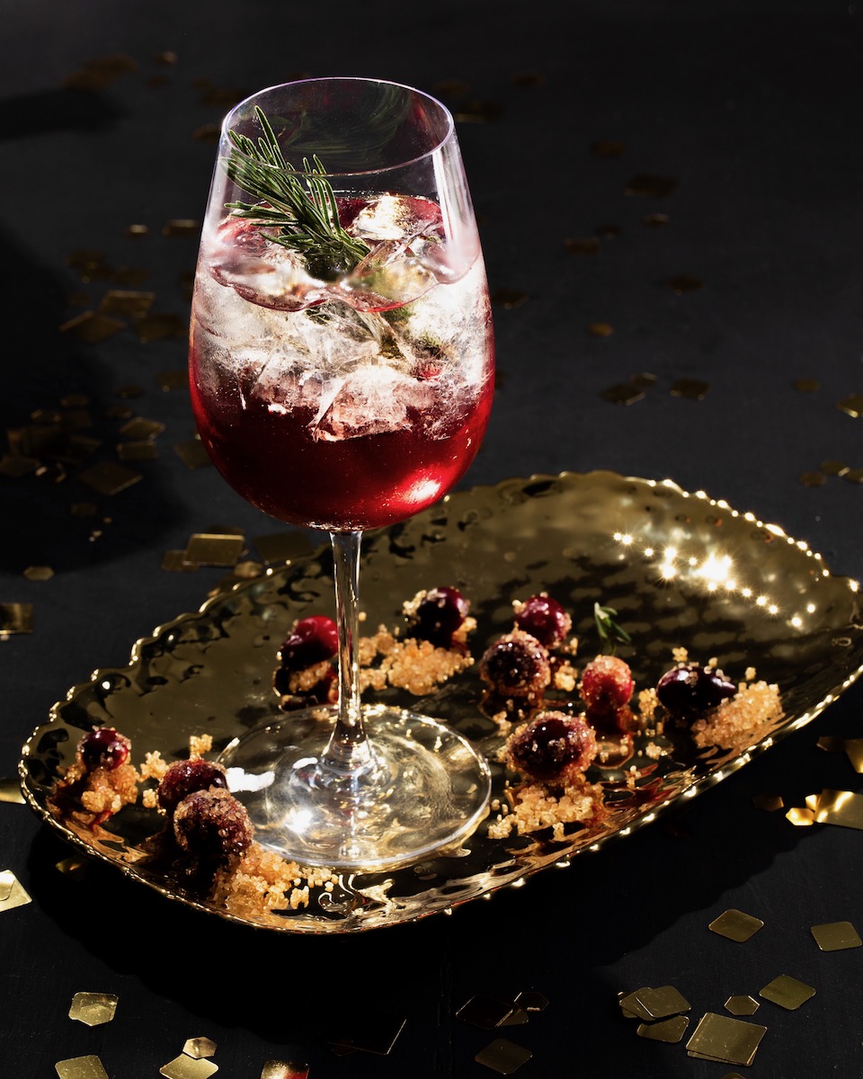 A rosemary cocktail in a white wine glass, gleaming with bright red cranberry juice and staged on a black background
