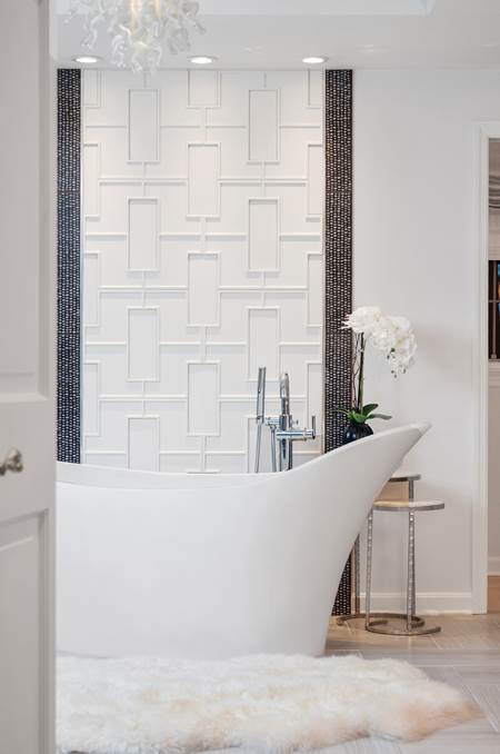 A white free standing tub with a white fur carpet and detailed white tile wall in a bathroom designed by Notion Design. 