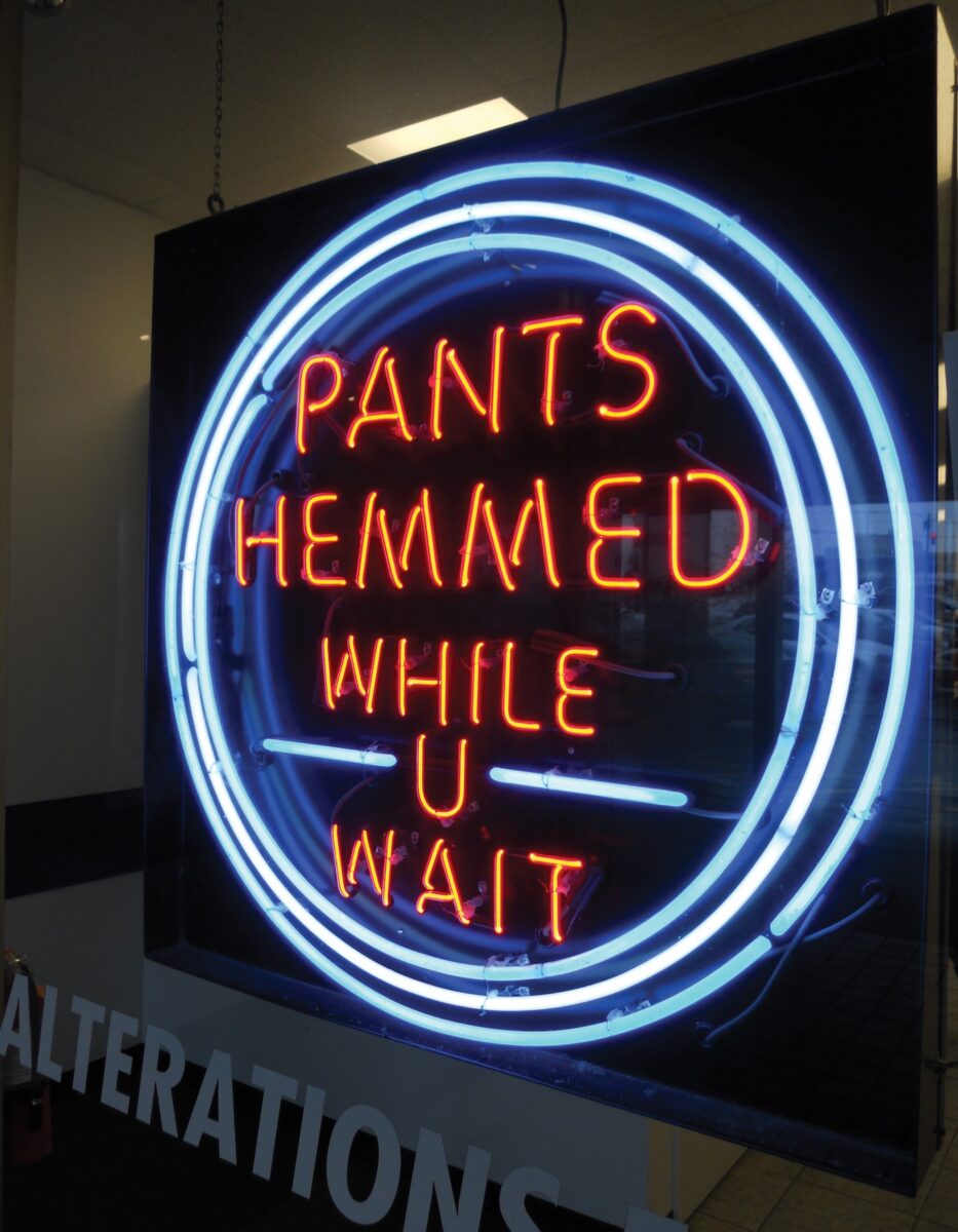 A red neon sign with blue circles around it that says Pants Hemmed While U Wait.