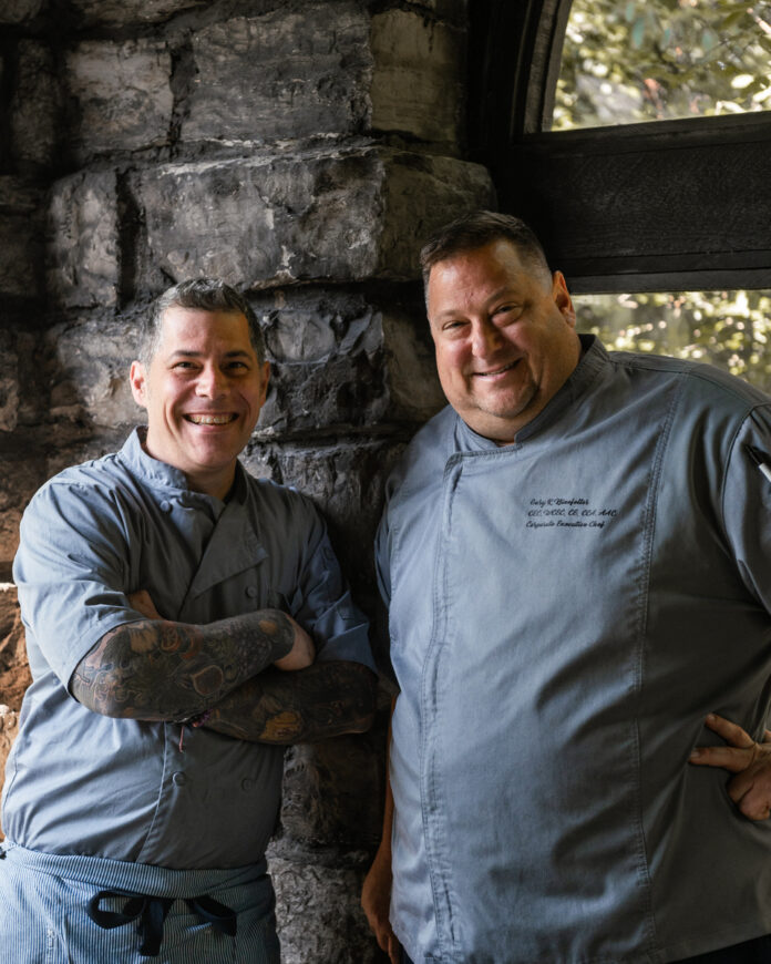Two professional chefs posing in a kitchen, one with tattoos, preparing for an Italian-American Feast of the Seven Fishes tradition.