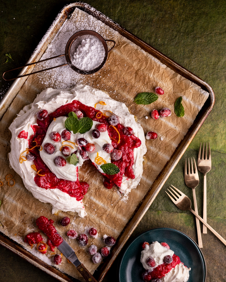 A large meringue topped with whipped cream, fresh cranberries, orange zest and mint leaves sits on a parchment lined baking tray.