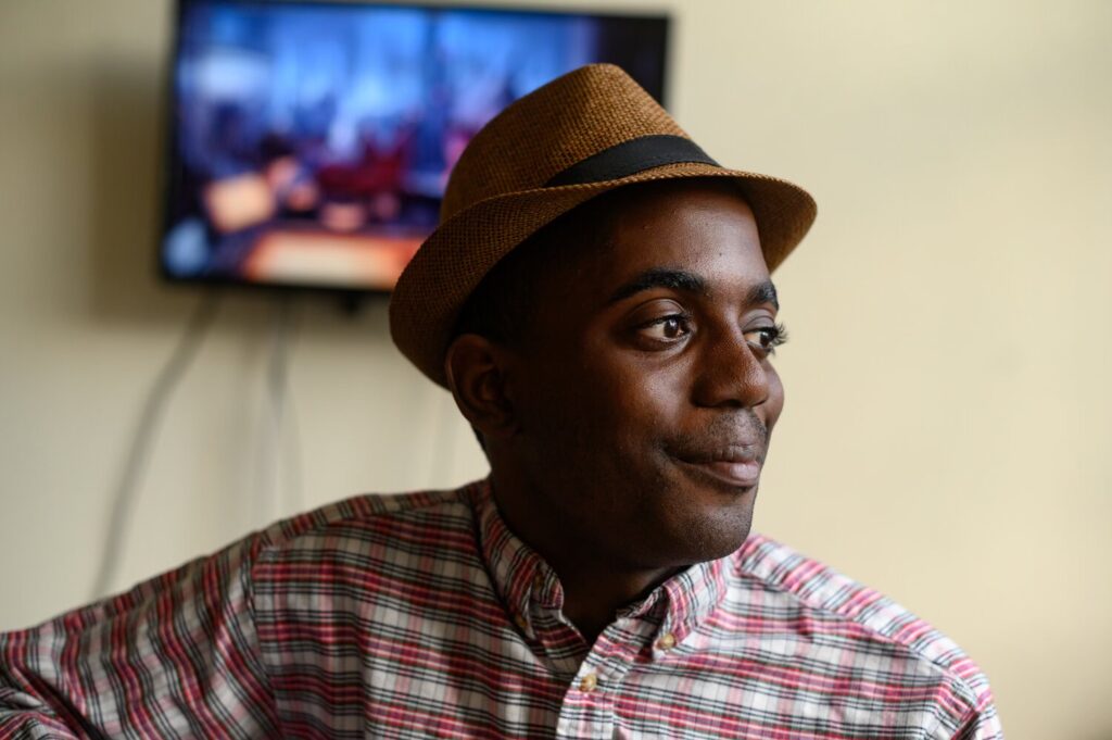 A man from ACTION Housing Pittsburgh looks off to the right side with a brown fedora and a red plaid shirt on.