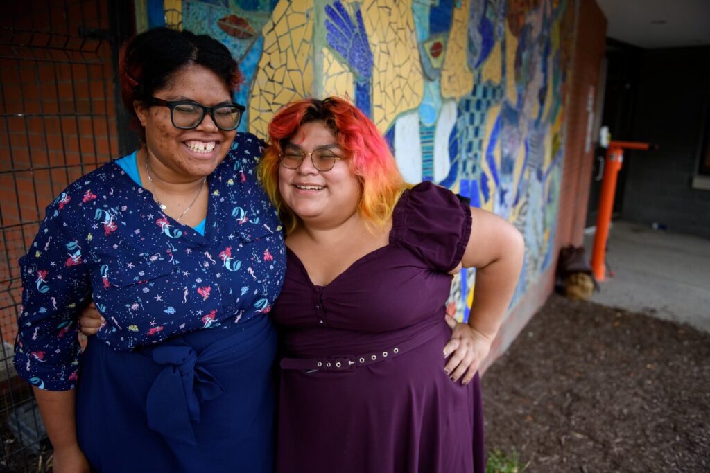 A woman with orange and pink hair and a purple dress stands with an arm around a woman in a blue dress with a graffiti wall behind them.