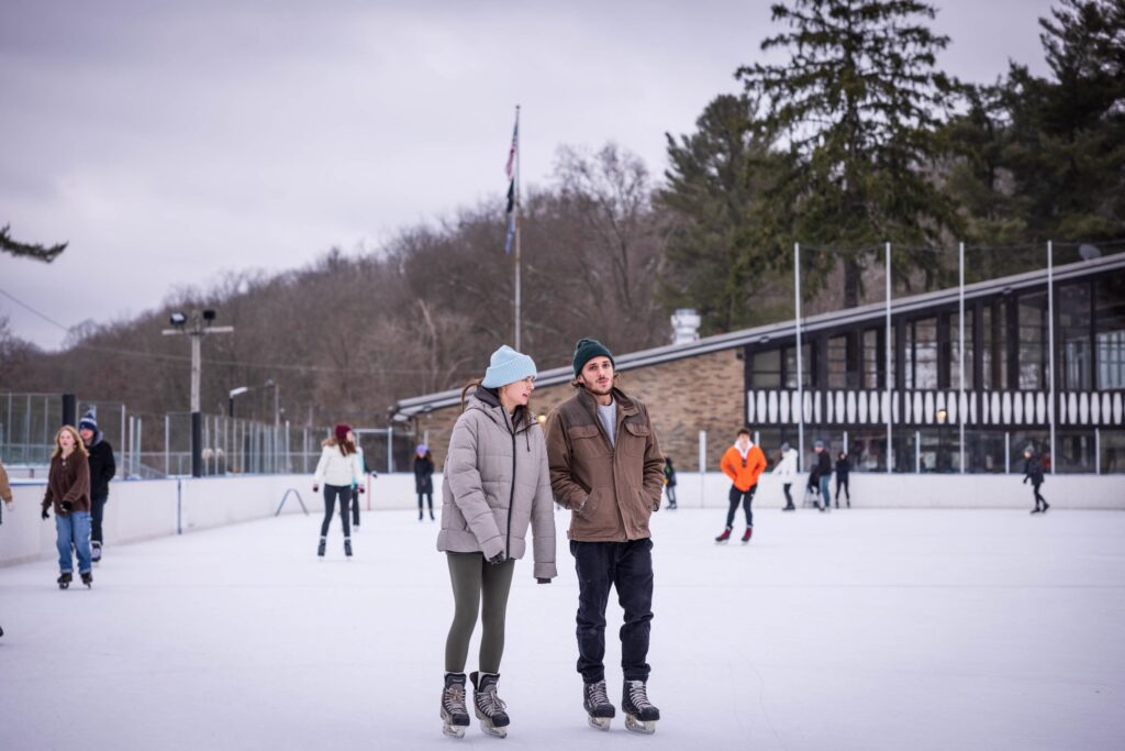 A man and women with hats and winter coats, skating at an outdoor skating rink.