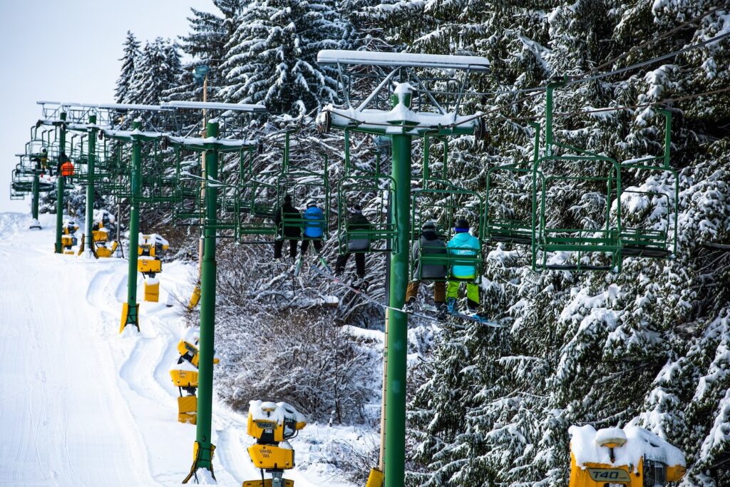 A picture of a ski slope chair lift next to a row of evergreen trees with people riding in the lift chairs on a snow covered day.